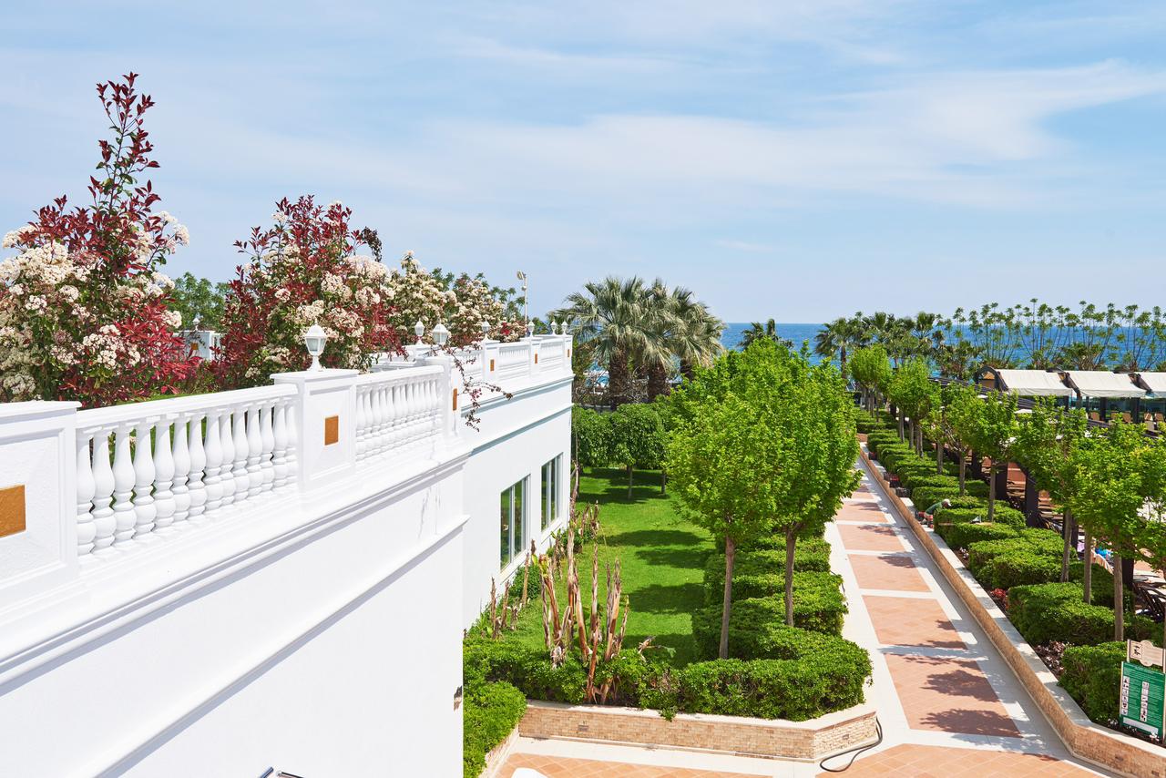 A well-maintained garden with green trees, a white building with a decorative railing, and a pathway leading towards the ocean in the background.
